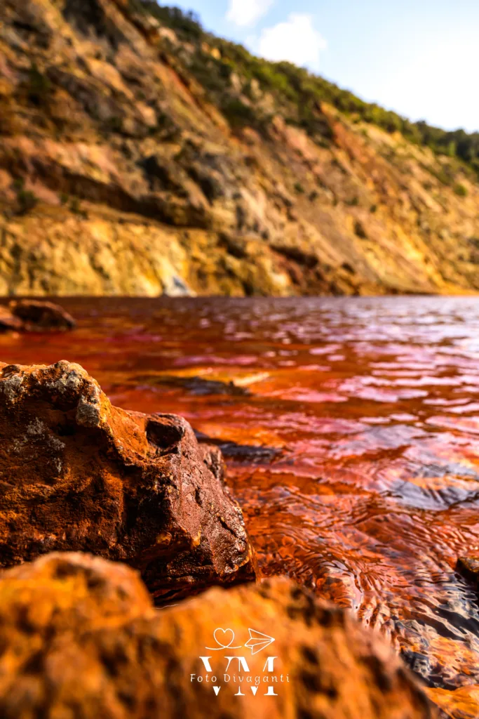 Rocce arancioni vicino a un corso d'acqua rossastra con collina rocciosa sullo sfondo.