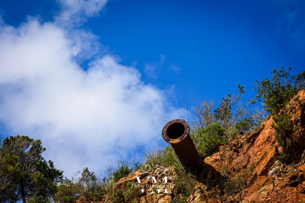 Tubo arrugginito che sporge da parete rocciosa con vegetazione e cielo azzurro parzialmente nuvoloso.