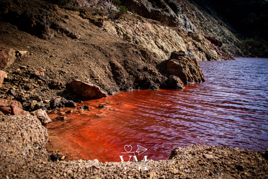 Lago con acqua rossa intensa su riva rocciosa con scogliere marroni e beige.