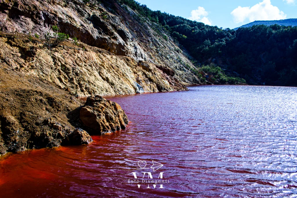 Lago con acqua rossa circondato da rocce e vegetazione sotto cielo parzialmente nuvoloso.
