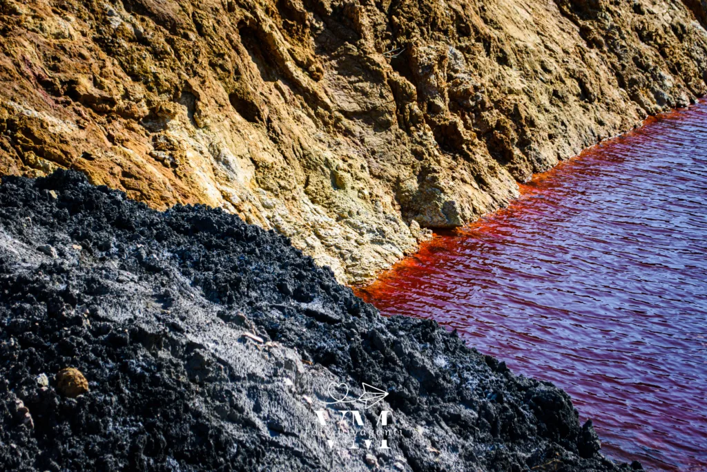Riva rocciosa con rocce nere e giallo-marroni accanto a un corpo d'acqua rosso intenso.