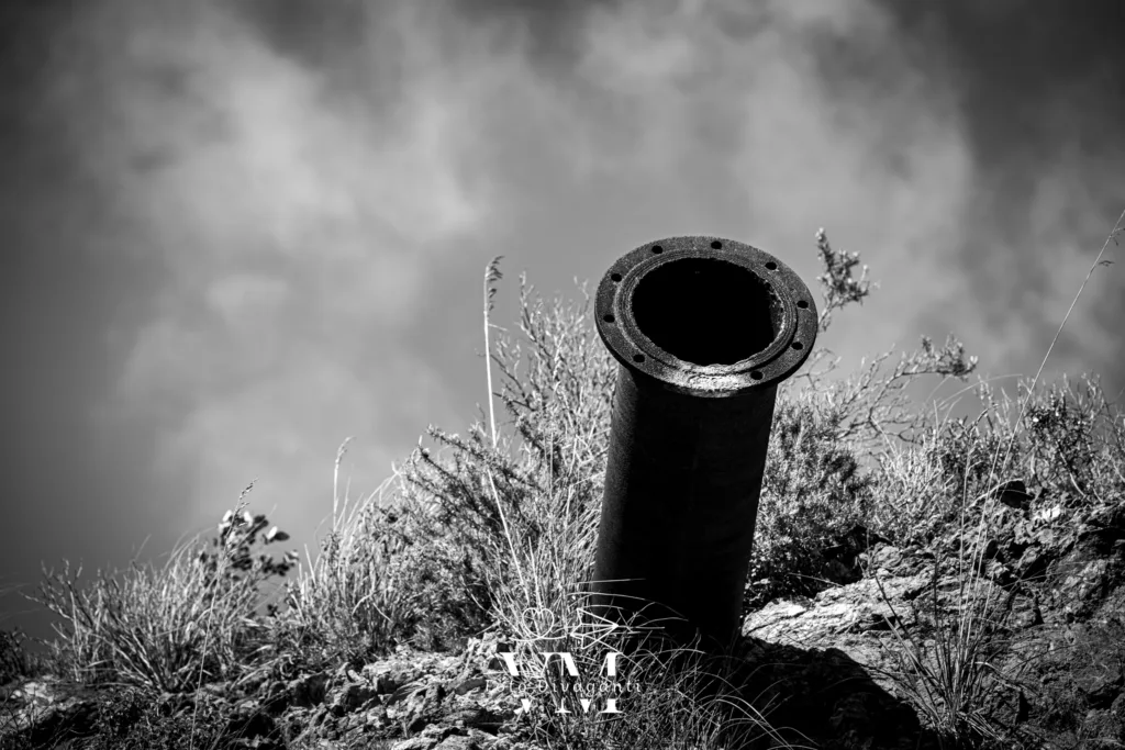 Tubo metallico arrugginito che sporge da terreno roccioso con erbe sotto un cielo nuvoloso.