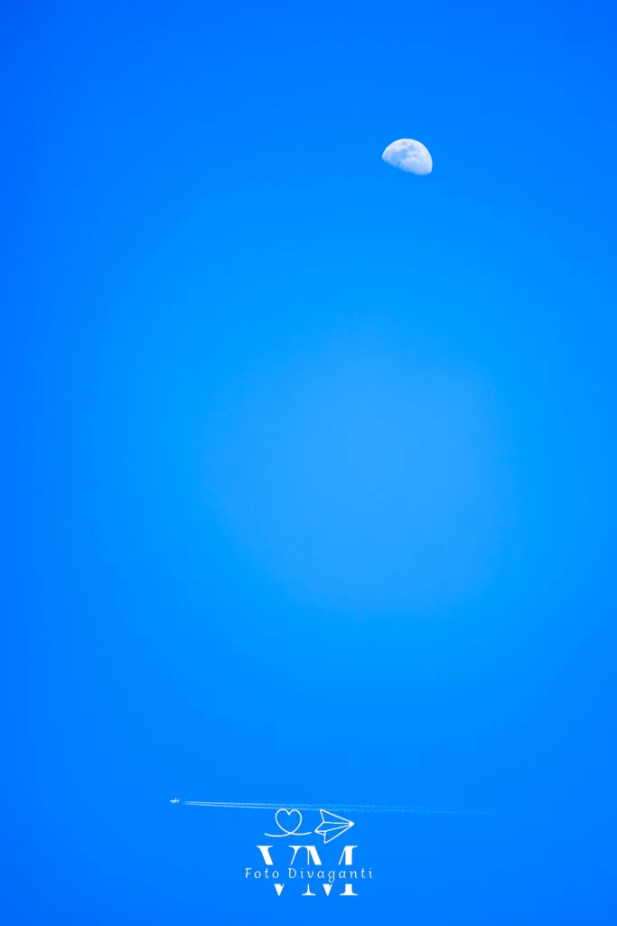 Luna parziale in cielo blu con aereo e scia di condensazione in basso a sinistra.